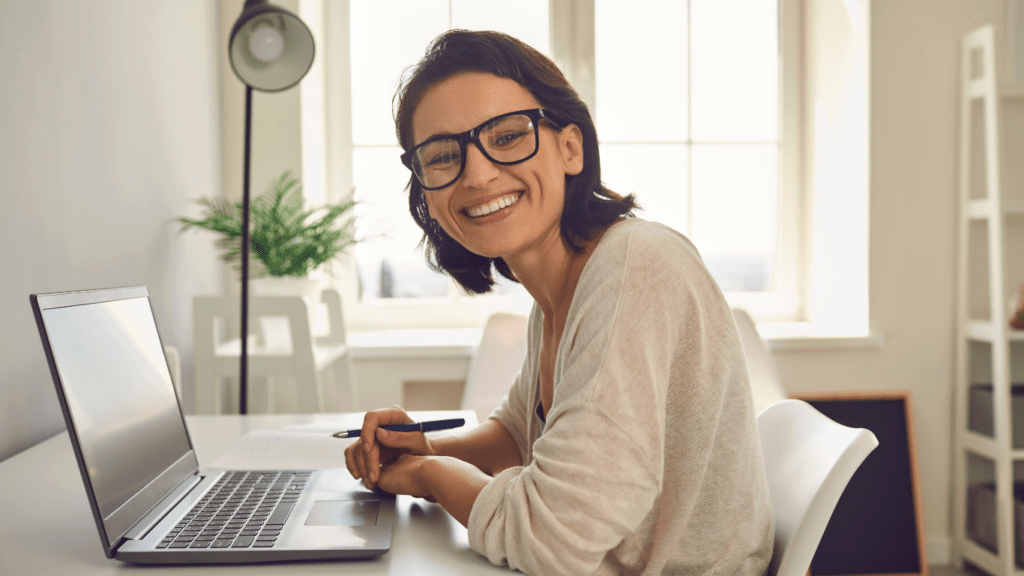 A smiling person with glasses sitting at a desk with a laptop