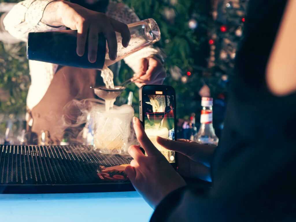 A bartender expertly prepares a cocktail while a patron captures the moment with a smartphone in a lively bar.