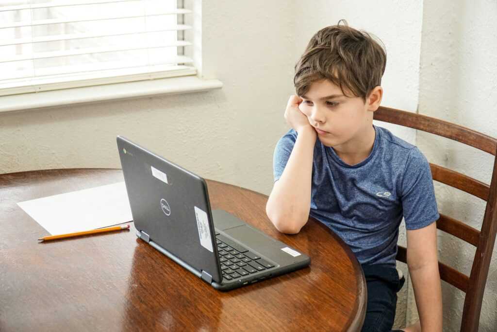 A young boy sitting at the kitchen table and looking at a laptop, bored. 