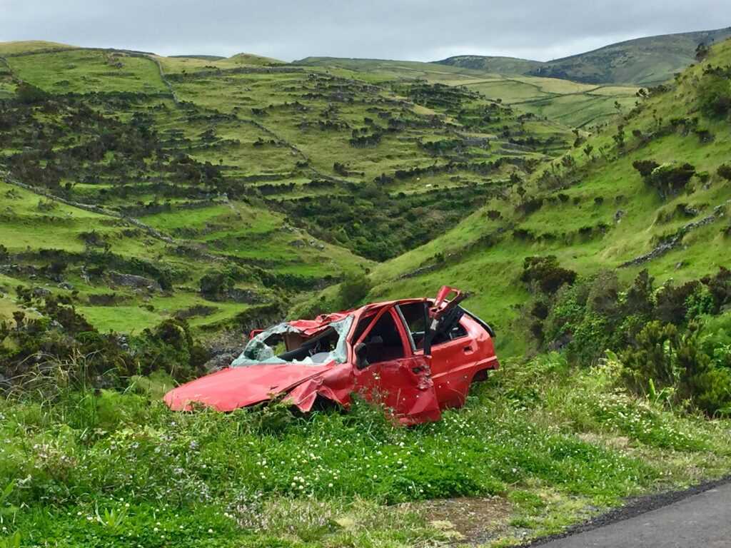 A red wrecked car lies abandoned in a scenic green valley, highlighting rural decay.